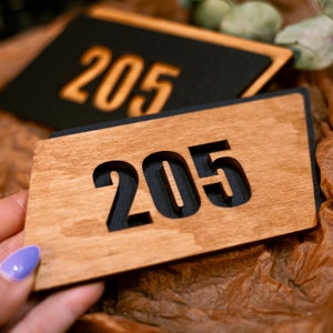 May include: A close-up of a wooden sign with the number 205 cut out and painted black. The sign is made of light brown wood and has a slightly rough texture.