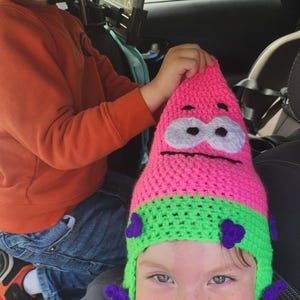 May include: A handmade crochet hat in the shape of Patrick Star from Spongebob Squarepants. The hat is primarily pink with a green brim and purple accents. The hat is being worn by a child.