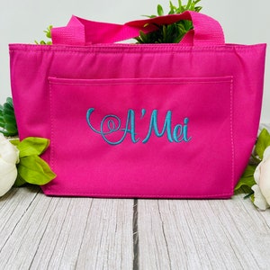 May include: A pink insulated lunch bag with a front pocket and a teal embroidered name "A'Mei".