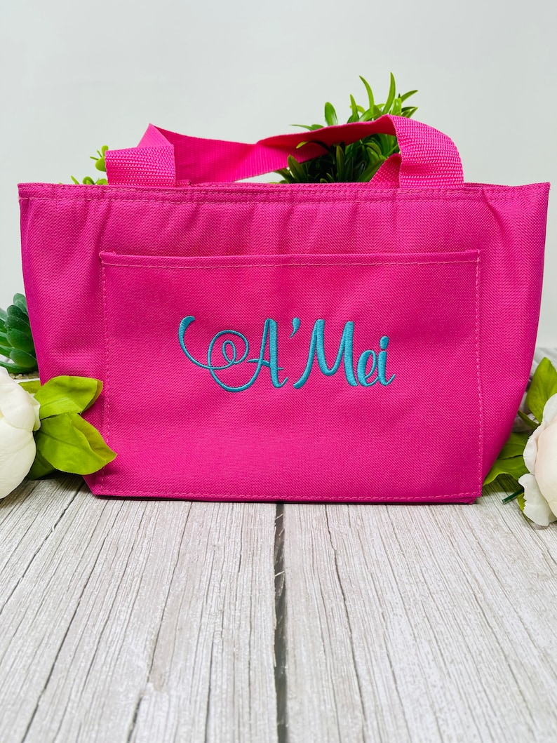 May include: A pink insulated lunch bag with a front pocket and a teal embroidered name "A'Mei".