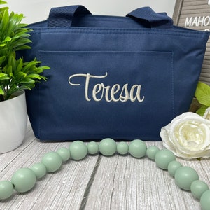 May include: A navy blue tote bag with a white embroidered name "Teresa" on the front. The bag is sitting on a wooden surface with a string of light green beads and a white flower.