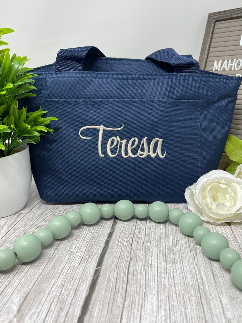 May include: A navy blue tote bag with a white embroidered name "Teresa" on the front. The bag is sitting on a wooden surface with a string of light green beads and a white flower.