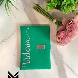 May include: A green leather bound journal with a silver clasp and the name "Victoria" written in pink script on the front.