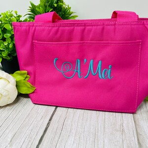 May include: A pink insulated lunch bag with a front pocket and two handles. The bag is embroidered with the name "A'Mei" in blue thread.