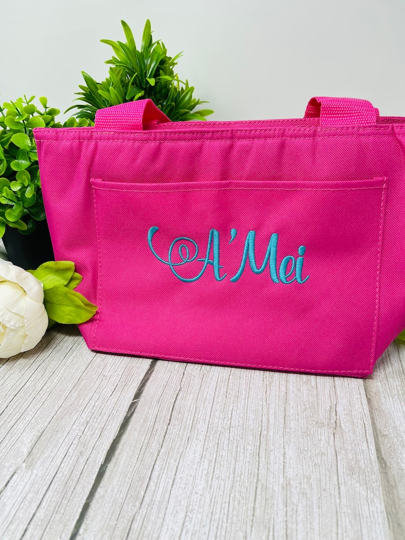 May include: A pink insulated lunch bag with a front pocket and two handles. The bag is embroidered with the name "A'Mei" in blue thread.