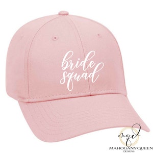 May include: Pink baseball cap with white cursive text that reads "bride squad".