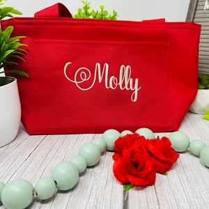 May include: Red insulated tote bag with a white embroidered name "Molly" on the front. The bag is sitting on a wooden surface with a string of light green beads and three red roses.