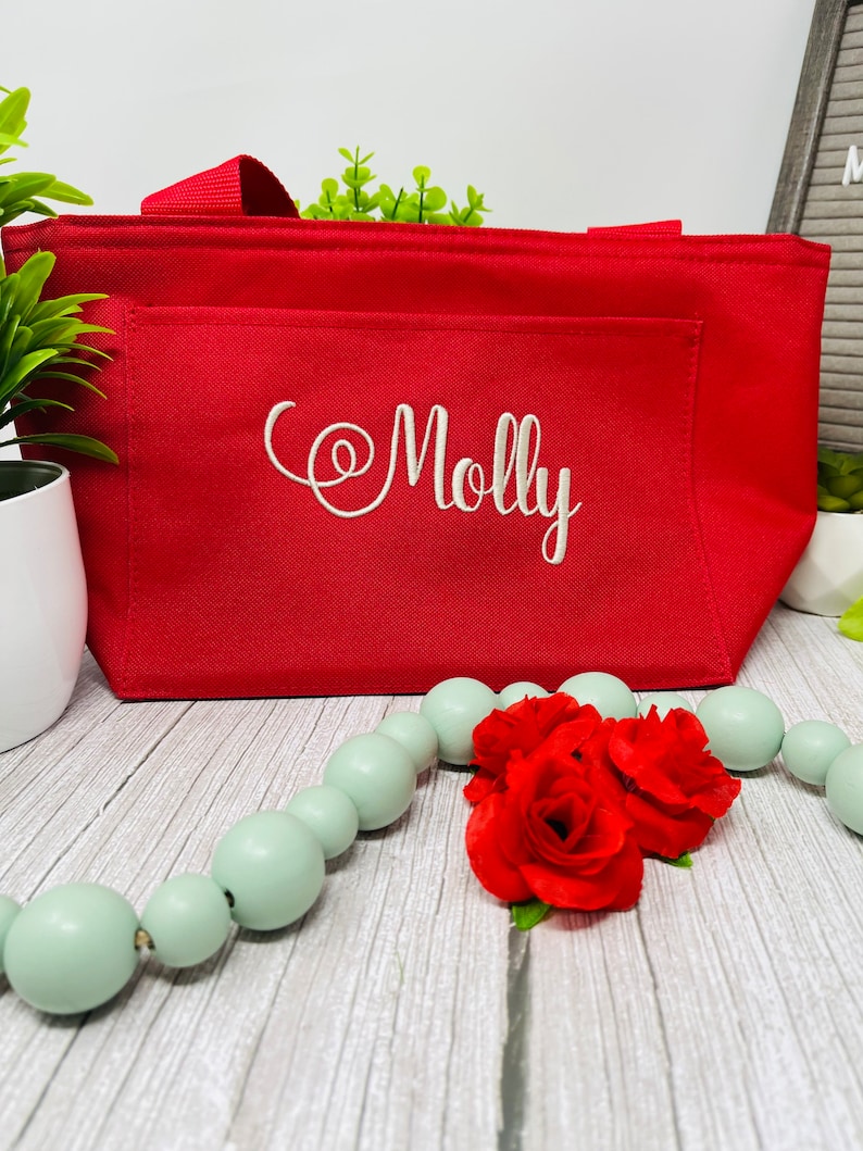 May include: Red insulated tote bag with a white embroidered name "Molly" on the front. The bag is sitting on a wooden surface with a string of light green beads and three red roses.