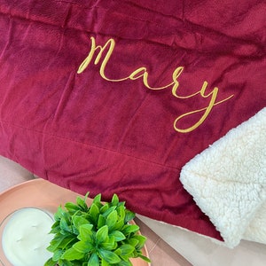 May include: A burgundy fleece blanket with a white sherpa lining. The blanket has the name "Mary" embroidered in gold thread.