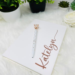 May include: White notebook with the name "Katelyn" written in gold script. A white pen with gold accents and a clear diamond-shaped top is on the notebook.
