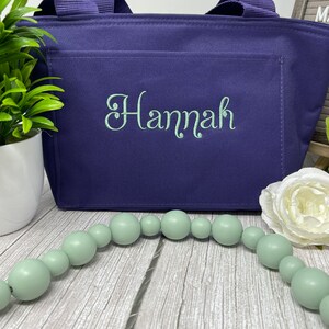 May include: A purple tote bag with a zippered top and a front pocket. The bag has the name "Hannah" embroidered on the front pocket in a light green color.