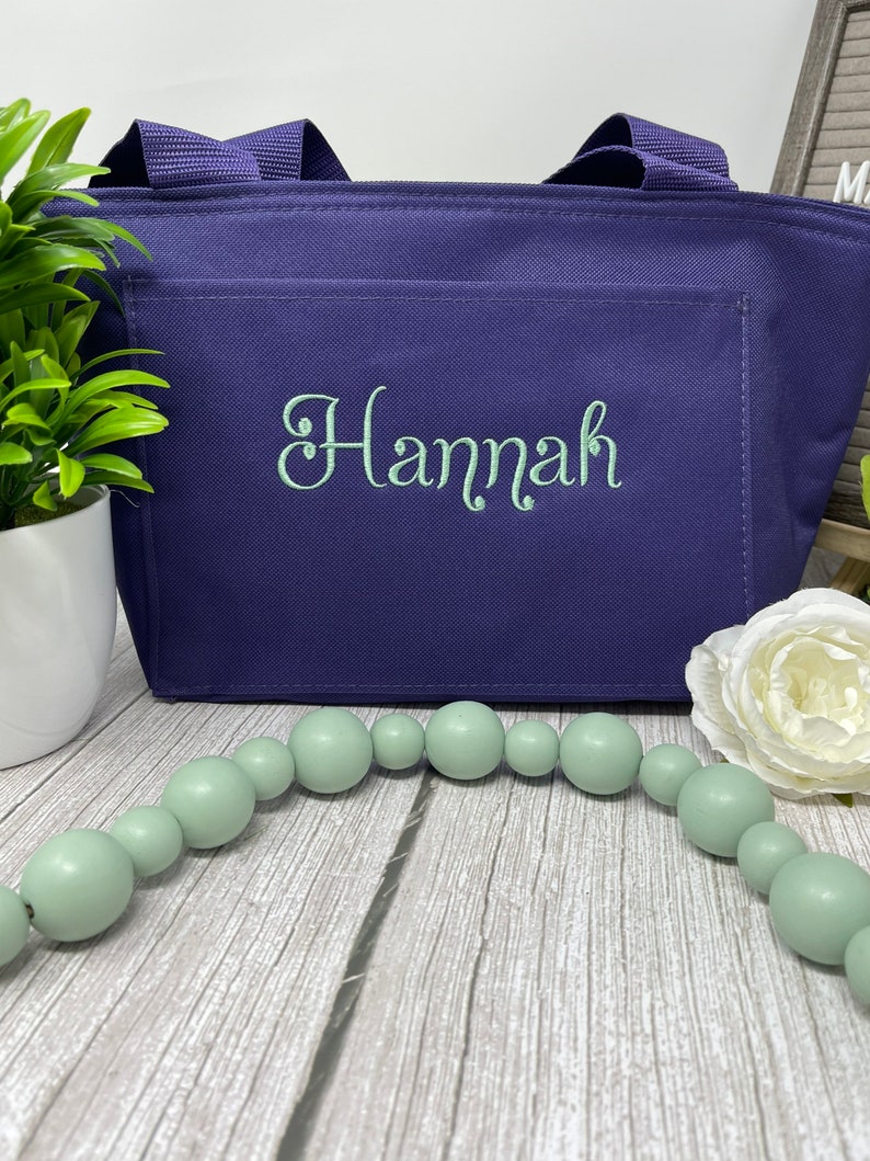 May include: A purple tote bag with a zippered top and a front pocket. The bag has the name "Hannah" embroidered on the front pocket in a light green color.