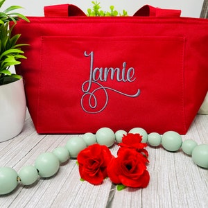 May include: Red insulated lunch bag with a zipper closure and a front pocket. The bag has the name "Jamie" embroidered on the front in gray thread.