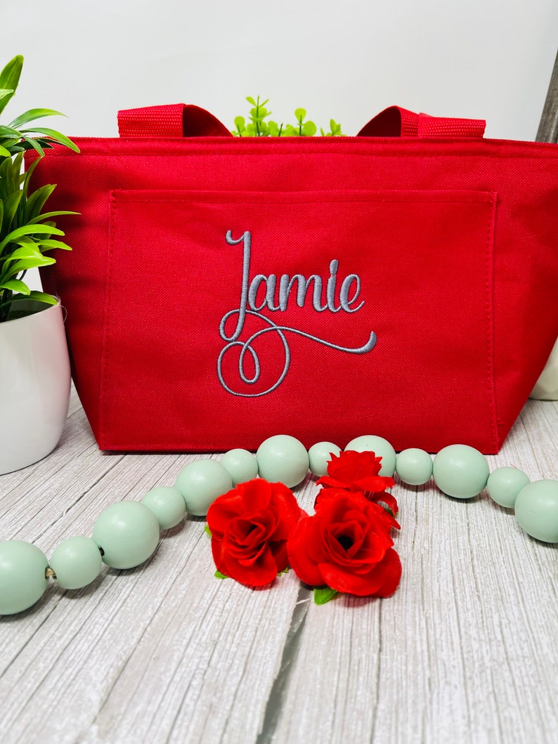 May include: Red insulated lunch bag with a zipper closure and a front pocket. The bag has the name "Jamie" embroidered on the front in gray thread.