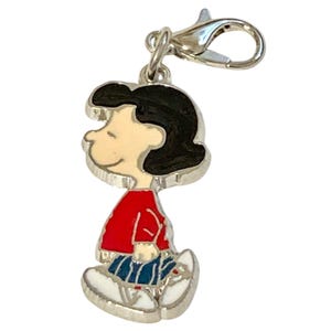 May include: A silver charm depicting a cartoon character with black hair, a red top, blue skirt, and white shoes. The charm has a lobster clasp for attaching to a bracelet or necklace. The character is smiling.