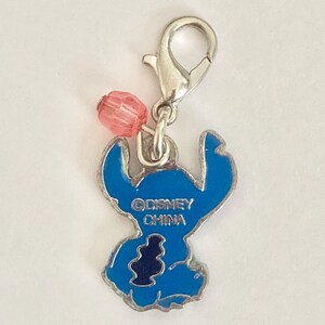 Puede incluir: Un charm de esmalte azul con el personaje Stitch de Lilo & Stitch de Disney. El charm tiene un cierre plateado y una cuenta rosa. El texto "&copy;DISNEY CHINA" es visible en el charm.