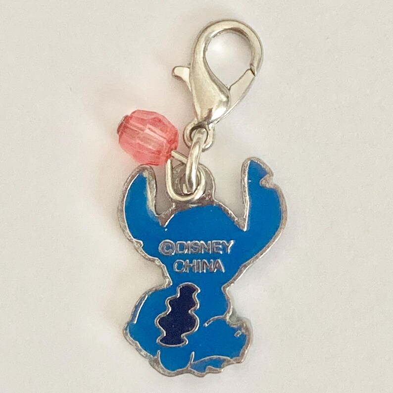 Puede incluir: Un charm de esmalte azul con el personaje Stitch de Lilo & Stitch de Disney. El charm tiene un cierre plateado y una cuenta rosa. El texto "&copy;DISNEY CHINA" es visible en el charm.