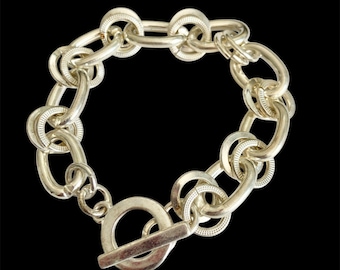 May include: Silver-toned chain bracelet with a toggle clasp. The bracelet features a series of oval and circular links, some with textured detailing. The background is solid black, highlighting the jewelry.