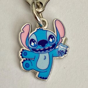 Puede incluir: Un dije de esmalte azul y rosa con el personaje Stitch de Lilo & Stitch. Stitch sostiene una bebida de t&eacute; boba. El dije est&aacute; unido a un cierre de metal plateado.