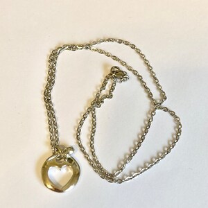 May include: A silver chain necklace with a heart-shaped pendant. The pendant is a circle with a heart cut out of the center.