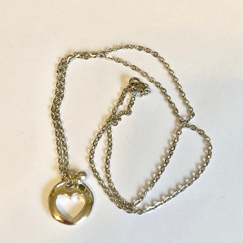 May include: A silver chain necklace with a heart-shaped pendant. The pendant is a circle with a heart cut out of the center.