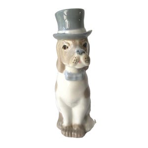 May include: A ceramic dog figurine with a gray top hat and a blue bow tie. The dog has brown and white fur, with brown spots on its body. The figurine is sitting upright, with its paws together.