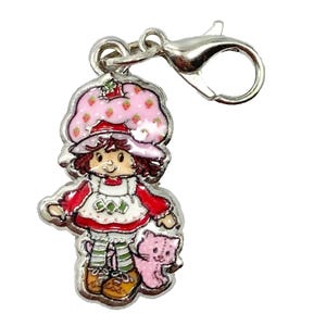 May include: A silver charm depicting a cartoon character with a strawberry hat, red dress, and a pink cat. The charm has a lobster clasp for attachment. The character is smiling and holding a pink cat. The charm is suitable for a bracelet or necklace.