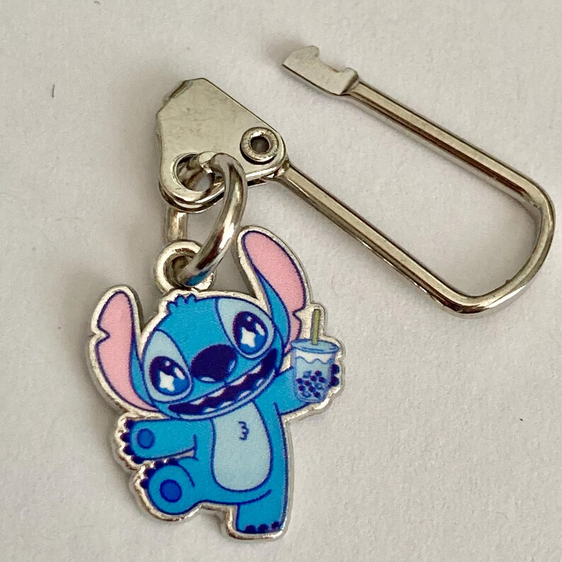 Puede incluir: Un dije de personaje de dibujos animados azul y rosa sosteniendo una bebida con una pajita. El dije est&aacute; unido a un cierre de metal plateado. El personaje tiene ojos grandes y una expresi&oacute;n sonriente. El dije es probablemente para un llavero o un tirador de cremallera.