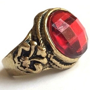 May include: A gold ring with a red gemstone. The ring has a detailed design with a floral motif.