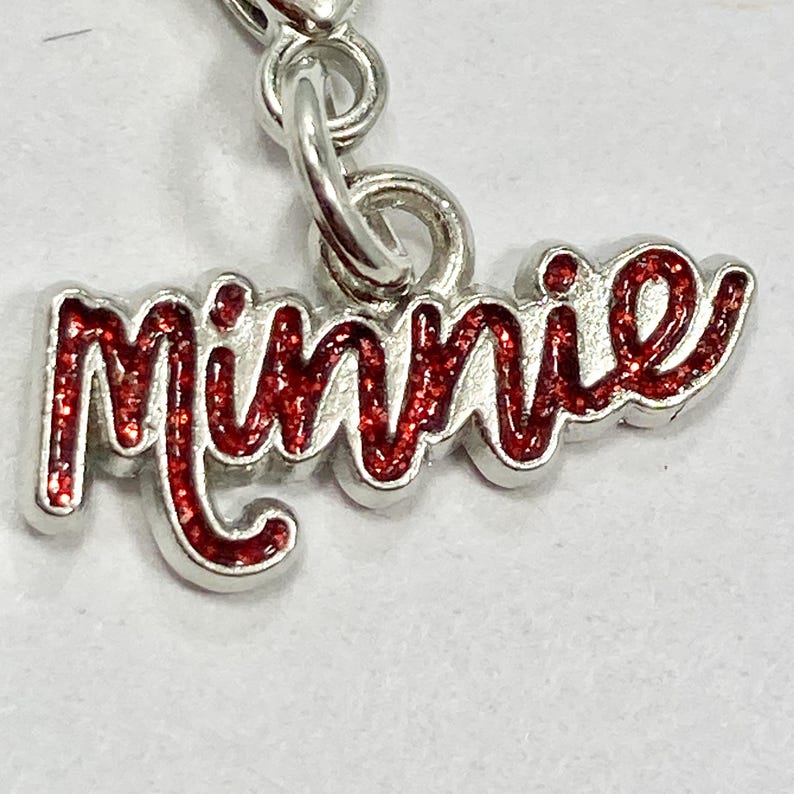 以下が含まれることがあります： 赤いグリッター文字で「Minnie」と書かれたシルバーのチャーム。チャームはシルバーのループに付いており、ペンダントやブレスレットやネックレスのチャームとして使用できます。デザインはシンプルでエレガントです。