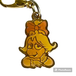 May include: Gold-colored metal keychain charm featuring a cartoon character with yellow hair, a pink bow, and a yellow and orange face.