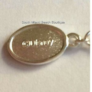 Puede incluir: Primer plano de una etiqueta ovalada de plata con la palabra "GUESS" grabada en ella.