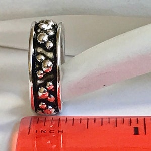 Puede incluir: Un anillo plateado con un dise&ntilde;o texturizado de formas redondeadas en relieve. El anillo est&aacute; junto a una regla. El dise&ntilde;o del anillo es oscuro, con un borde plateado brillante. El anillo mide aproximadamente 1,27 cm de ancho.