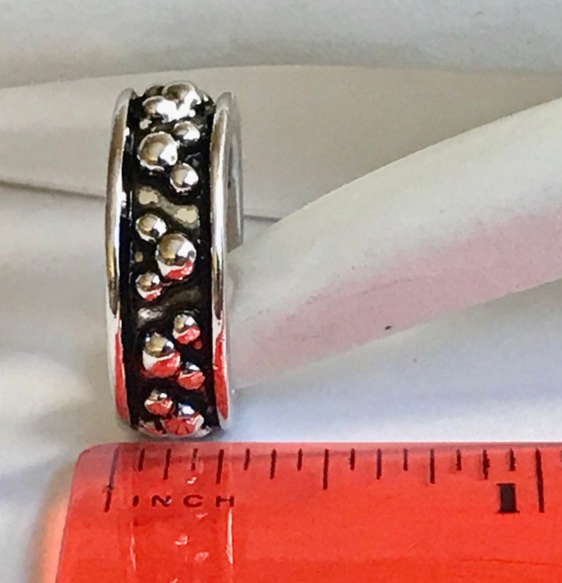 Puede incluir: Un anillo plateado con un dise&ntilde;o texturizado de formas redondeadas en relieve. El anillo est&aacute; junto a una regla. El dise&ntilde;o del anillo es oscuro, con un borde plateado brillante. El anillo mide aproximadamente 1,27 cm de ancho.