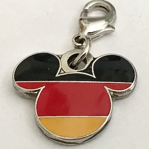 May include: A silver charm in the shape of Mickey Mouse's head with the colors of the German flag. The charm has a black top, a red middle, and a yellow bottom. The charm has a silver lobster clasp.