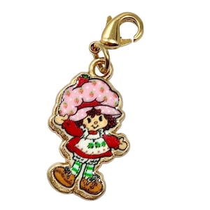 May include: A gold-coloured charm depicting a cartoon character with a pink strawberry hat, red dress, and green and white striped socks. The charm has a lobster clasp for attachment to a bracelet or necklace.