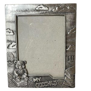 May include: A silver-colored picture frame with a teddy bear, clouds, and the words "MY GODCHILD." The frame has a rectangular opening and is decorated with a bear reading a book and stacks of books.