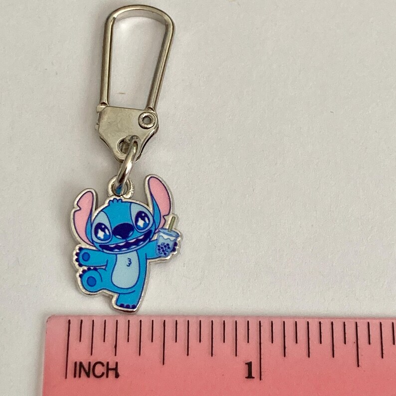 Puede incluir: Un charm de esmalte azul y rosa con el personaje Stitch de Lilo & Stitch, sosteniendo una bebida. El charm est&aacute; unido a un cierre de metal plateado. El charm mide aproximadamente 2,5 cm de largo.