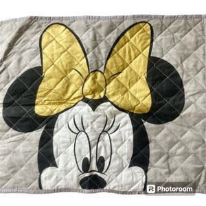 May include: A quilted fabric panel featuring a large graphic of Minnie Mouse. The design displays a yellow bow, black ears, and a white face with black outlines on a light gray background. The entire surface has a diamond-shaped stitched pattern.