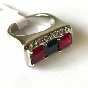 May include: A silver ring with a black and two red square-shaped stones set in a row. The ring is decorated with a row of clear rhinestones.