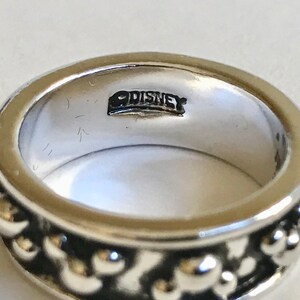 Puede incluir: Primer plano de un anillo plateado con un dise&ntilde;o de cuentas alrededor de la banda. El interior del anillo est&aacute; estampado con la palabra "DISNEY". El anillo es una joya sencilla pero elegante.