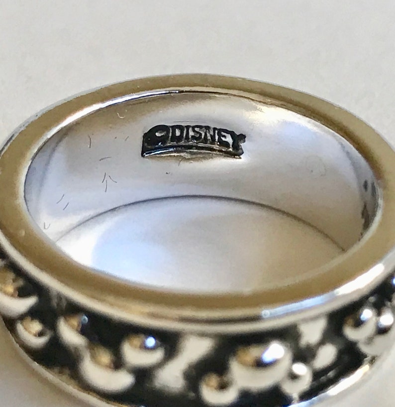 Puede incluir: Primer plano de un anillo plateado con un dise&ntilde;o de cuentas alrededor de la banda. El interior del anillo est&aacute; estampado con la palabra "DISNEY". El anillo es una joya sencilla pero elegante.