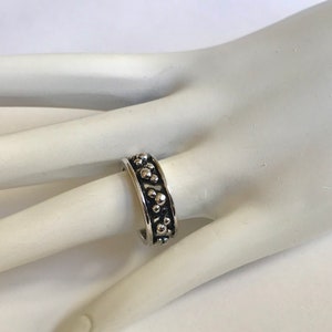 Puede incluir: Un anillo plateado con una banda texturizada adornada con peque&ntilde;os dise&ntilde;os circulares en relieve. El anillo se muestra en un dedo, resaltando su dise&ntilde;o detallado y acabado met&aacute;lico. El estilo del anillo sugiere un tema inspirado en la naturaleza.