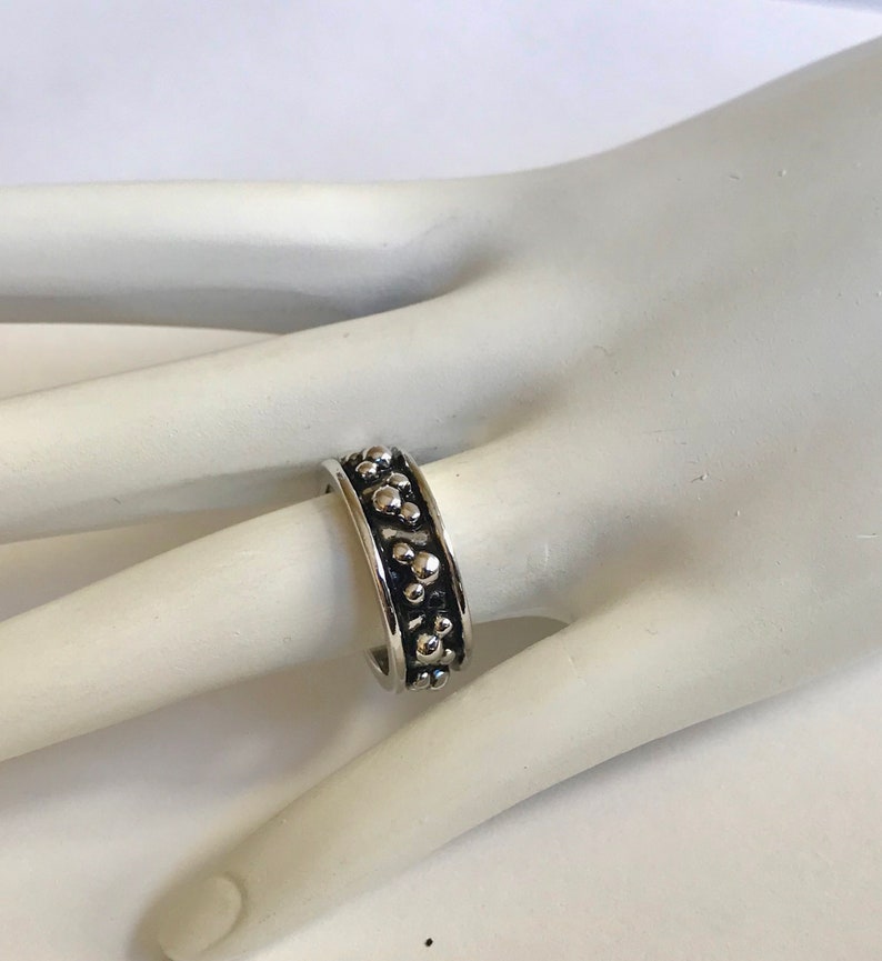 Puede incluir: Un anillo plateado con una banda texturizada adornada con peque&ntilde;os dise&ntilde;os circulares en relieve. El anillo se muestra en un dedo, resaltando su dise&ntilde;o detallado y acabado met&aacute;lico. El estilo del anillo sugiere un tema inspirado en la naturaleza.
