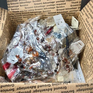 May include: A cardboard box filled with various jewelry items, including necklaces and earrings. The jewelry pieces are in clear plastic bags, with tags attached. The box has printed text on the sides.