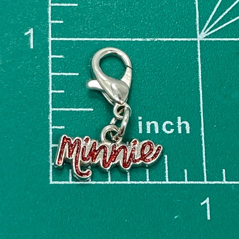 以下が含まれることがあります： 赤いグリッター文字で「Minnie」と書かれた、ロブスタークラスプ付きのシルバーチャーム。 チャームの長さは約2.5cmで、インチの目盛りが付いた緑色の背景に表示されています。