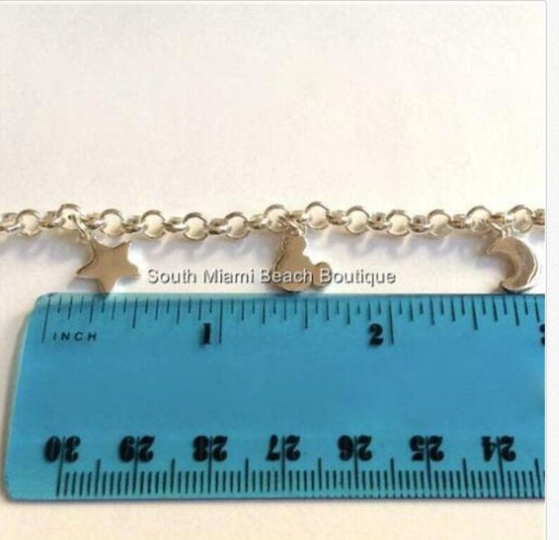 Puede incluir: Un brazalete de cadena de plata con dijes de estrella, luna y coraz&oacute;n. El brazalete se mide en una regla que muestra que mide poco m&aacute;s de 1 pulgada de largo.