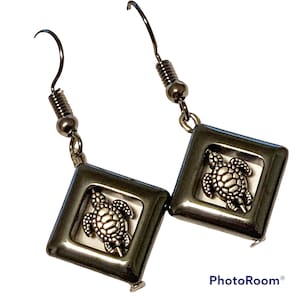 May include: A pair of silver earrings with a square-shaped pendant featuring a silver turtle design. The earrings have a silver hook fastening.