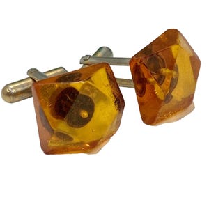 May include: A pair of cufflinks with a silver-colored backing and amber-colored, faceted, square-shaped faces. The amber faces have dark brown inclusions and a glossy finish. The cufflinks are set against a white background.