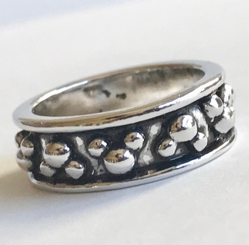 Puede incluir: Un anillo plateado con un dise&ntilde;o texturizado. El anillo presenta una banda de peque&ntilde;as formas esf&eacute;ricas en relieve alrededor de la circunferencia. El anillo tiene una superficie pulida y reflectante.